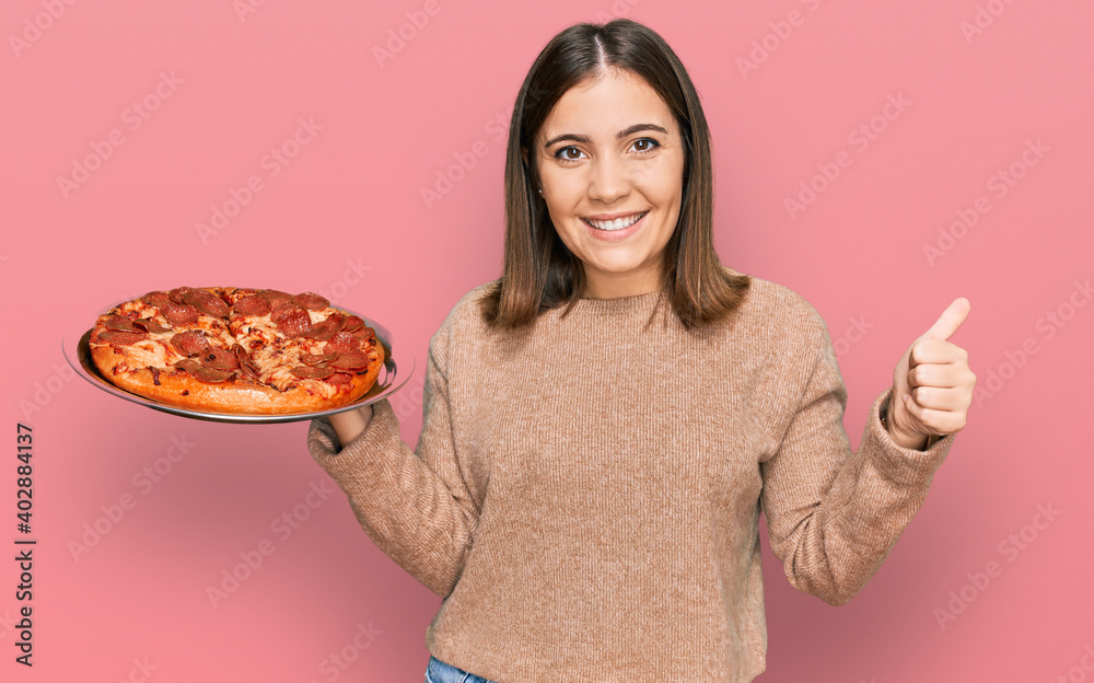 Young beautiful woman holding italian pizza smiling happy and positive, thumb up doing excellent and approval sign Young beautiful woman holding italian pizza smiling happy and positive, thumb up doing excellent and approval sign