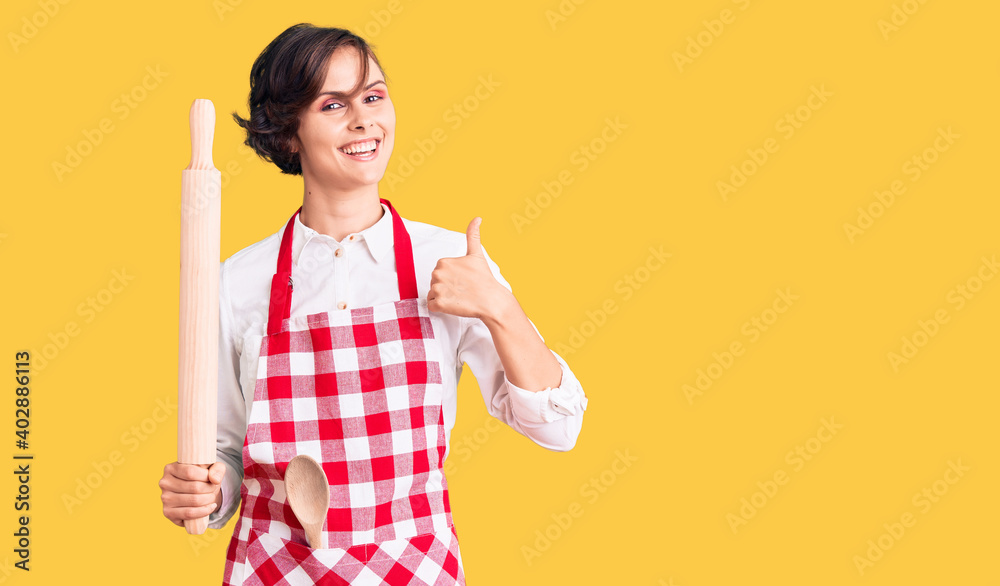Beautiful young woman with short hair wearing professional baker apron holding kneading roll smiling happy and positive, thumb up doing excellent and approval sign Beautiful young woman with short hair wearing professional baker apron holding kneading roll smiling happy and positive, thumb up doing excellent and approval sign