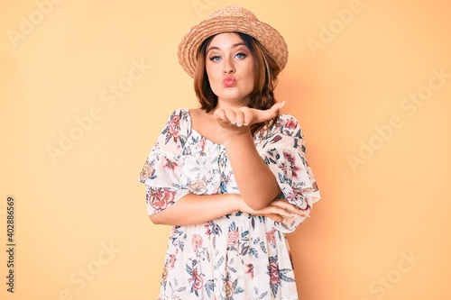 Young beautiful caucasian woman wearing summer dress and hat looking at the camera blowing a kiss with hand on air being lovely and sexy. love expression.