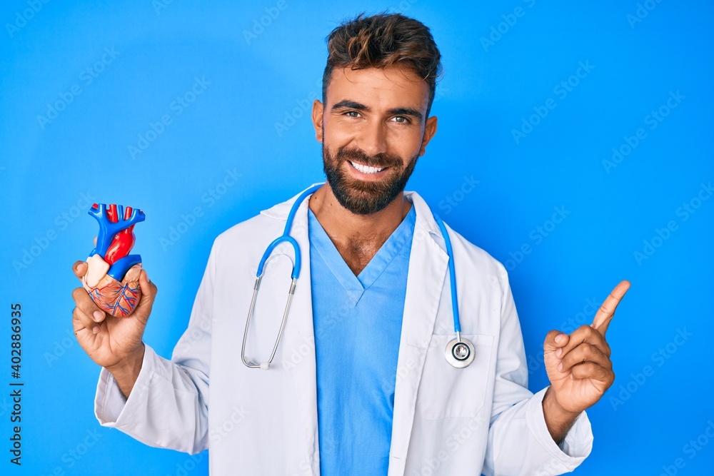 Young hispanic man wearing doctor uniform holding heart smiling happy pointing with hand and finger to the side Young hispanic man wearing doctor uniform holding heart smiling happy pointing with hand and finger to the side