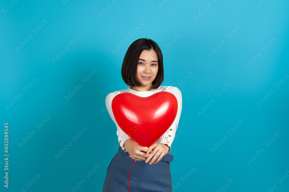 mock up a smiling Asian woman holds out a red heart-shaped balloon isolated on a blue background to the camera. mock up a smiling Asian woman holds out a red heart-shaped balloon isolated on a blue background to the camera.