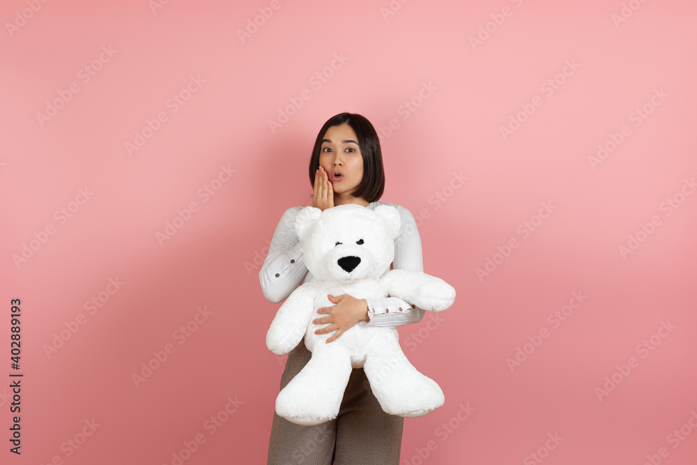 mock up surprised excited beautiful asian woman with open mouth and palm near face with white teddy bear isolated on pink background.