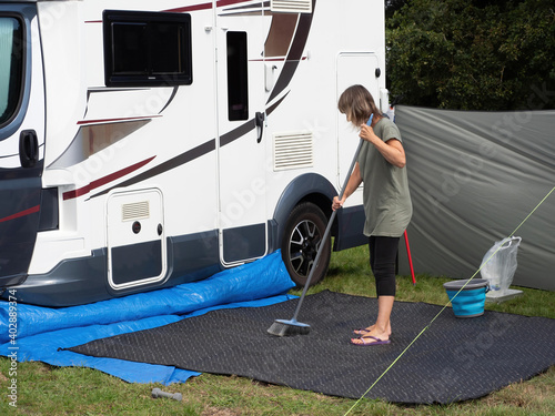 A lady motorhome owner sweeps her outdoor mat with a broom outside her recreational vehicle. A bucket and wind break are visible.