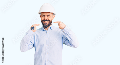 Young handsome man wearing architect hardhat smiling cheerful showing and pointing with fingers teeth and mouth. dental health concept.