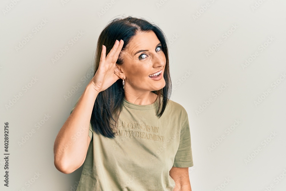 Middle age brunette woman wearing casual clothes smiling with hand over ear listening and hearing to rumor or gossip. deafness concept. Middle age brunette woman wearing casual clothes smiling with hand over ear listening and hearing to rumor or gossip. deafness concept.