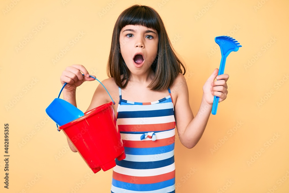 Young little girl with bang playing with summer shovel and bucket toys in shock face, looking skeptical and sarcastic, surprised with open mouth Young little girl with bang playing with summer shovel and bucket toys in shock face, looking skeptical and sarcastic, surprised with open mouth