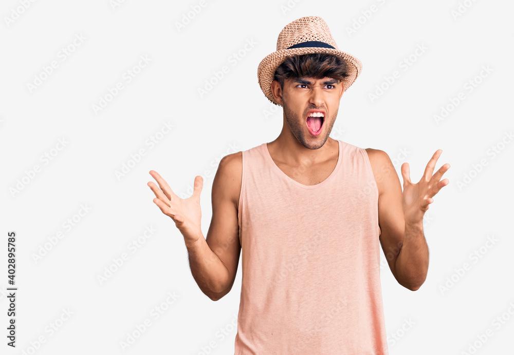 Young hispanic man wearing summer hat crazy and mad shouting and yelling with aggressive expression and arms raised. frustration concept. Young hispanic man wearing summer hat crazy and mad shouting and yelling with aggressive expression and arms raised. frustration concept.