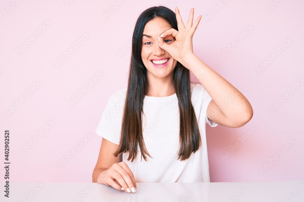 Young caucasian woman sitting at the table wearing casual white tshirt smiling happy doing ok sign with hand on eye looking through fingers Young caucasian woman sitting at the table wearing casual white tshirt smiling happy doing ok sign with hand on eye looking through fingers
