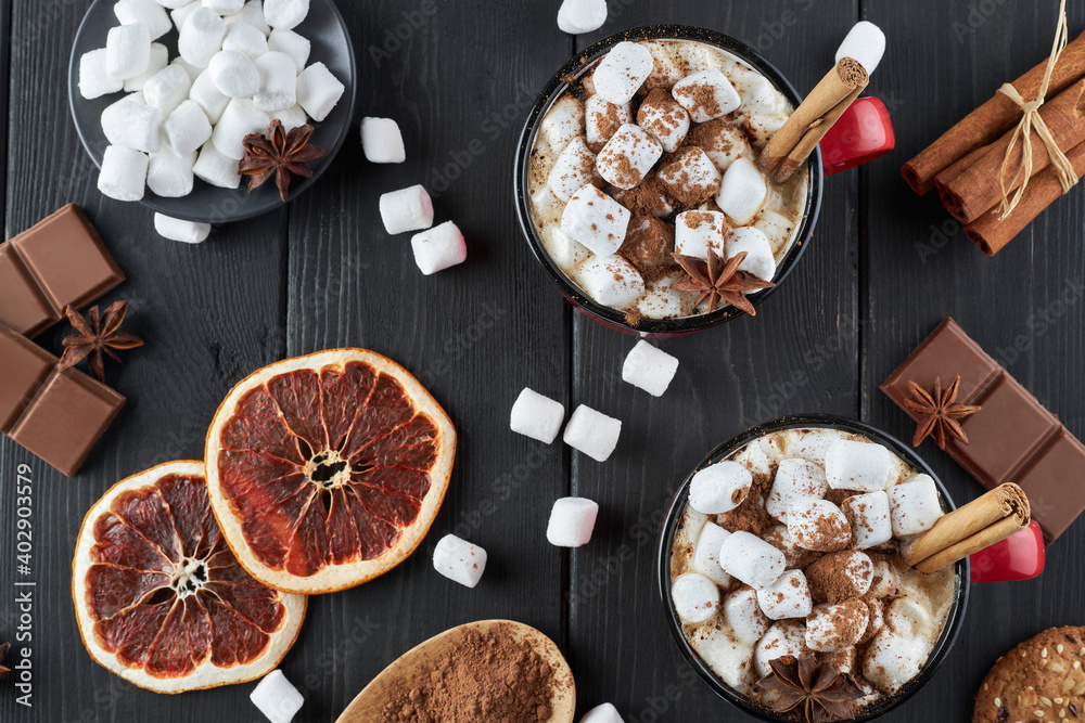 Fototapeta premium Two red cups of hot chocolate with marshmallow, anise and cinnamon sprinkled with cocoa powder on a wooden table with slices of dried grapefruit and chocolate pieces. Winter hot drink top view.