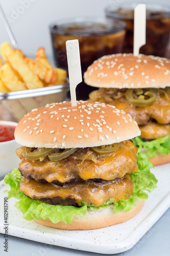 Double cheese burger with caramelized onion, lettuce and jalapeno, served with french fries and soda, on white plate, vertical