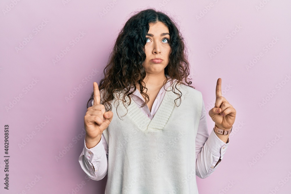 Young brunette woman with curly hair wearing casual clothes pointing up looking sad and upset, indicating direction with fingers, unhappy and depressed. Young brunette woman with curly hair wearing casual clothes pointing up looking sad and upset, indicating direction with fingers, unhappy and depressed.