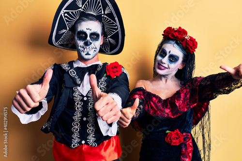 Young couple wearing mexican day of the dead costume over yellow approving doing positive gesture with hand, thumbs up smiling and happy for success. winner gesture.