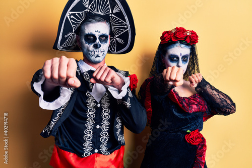 Young couple wearing mexican day of the dead costume over yellow punching fist to fight, aggressive and angry attack, threat and violence