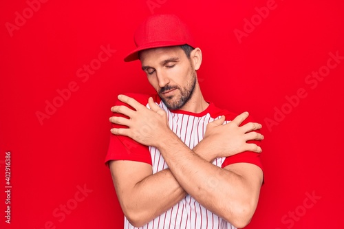 Young handsome player man wearing baseball sportswear over isolated red background hugging oneself happy and positive, smiling confident. Self love and self care © Krakenimages.com