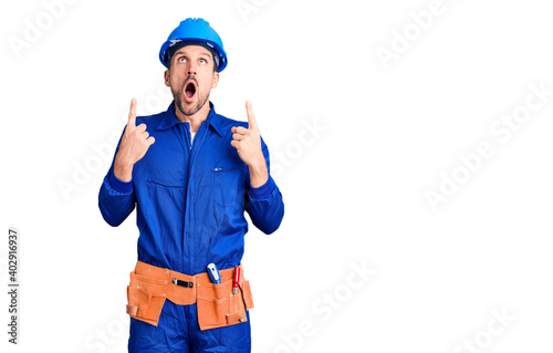 Young handsome man wearing worker uniform and hardhat looking fascinated with disbelief, surprise and amazed expression with hands on chin