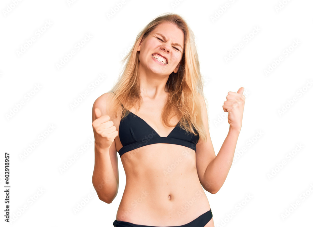 Young beautiful blonde woman wearing bikini very happy and excited doing winner gesture with arms raised, smiling and screaming for success. celebration concept.