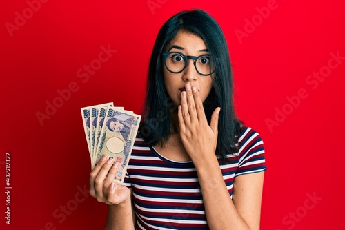 Beautiful asian young woman holding 5000 japanese yen banknotes covering mouth with hand, shocked and afraid for mistake. surprised expression