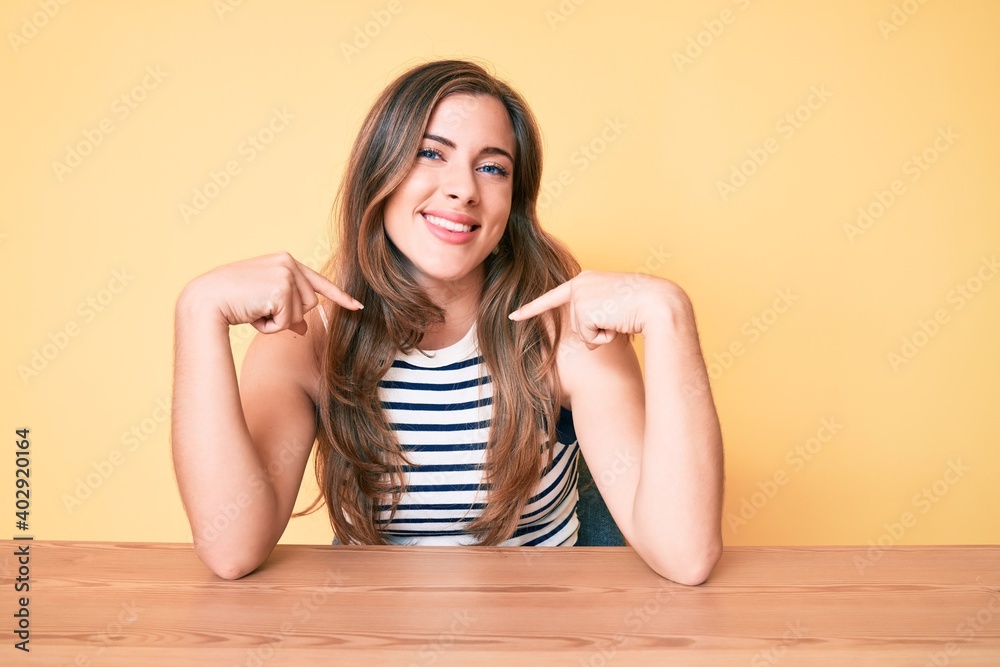 Beautiful young caucasian woman wearing casual clothes sitting on the table looking confident with smile on face, pointing oneself with fingers proud and happy. Beautiful young caucasian woman wearing casual clothes sitting on the table looking confident with smile on face, pointing oneself with fingers proud and happy.