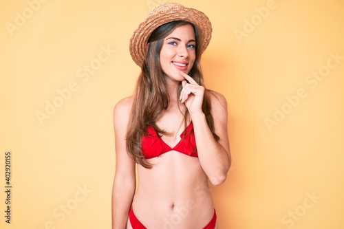 Beautiful young caucasian woman wearing bikini and hat smiling looking confident at the camera with crossed arms and hand on chin. thinking positive.