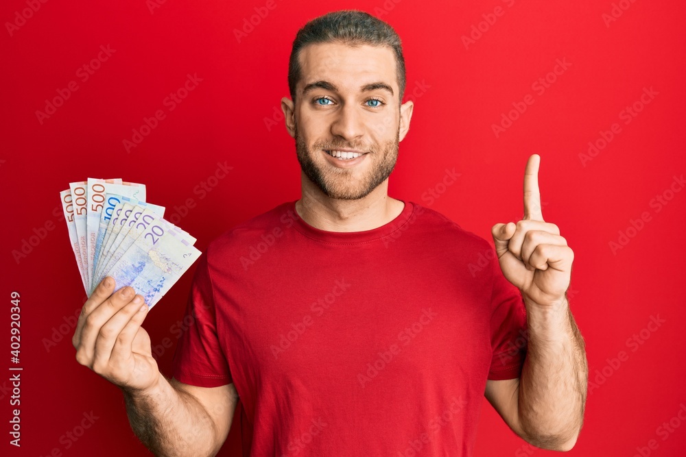 Young caucasian man holding swedish krona banknotes smiling with an idea or question pointing finger with happy face, number one Young caucasian man holding swedish krona banknotes smiling with an idea or question pointing finger with happy face, number one