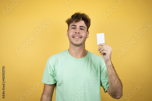 Handsome man wearing a green casual t-shirt over yellow background smiling and holding white card