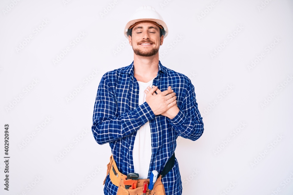 Young handsome man weaing handyman uniform smiling with hands on chest with closed eyes and grateful gesture on face. health concept. Young handsome man weaing handyman uniform smiling with hands on chest with closed eyes and grateful gesture on face. health concept.