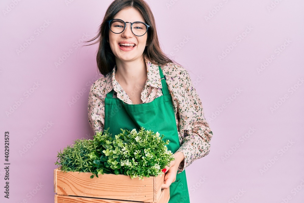 Young beautiful caucasian girl wearing gardener apron holding wooden plant pot smiling and laughing hard out loud because funny crazy joke. Young beautiful caucasian girl wearing gardener apron holding wooden plant pot smiling and laughing hard out loud because funny crazy joke.