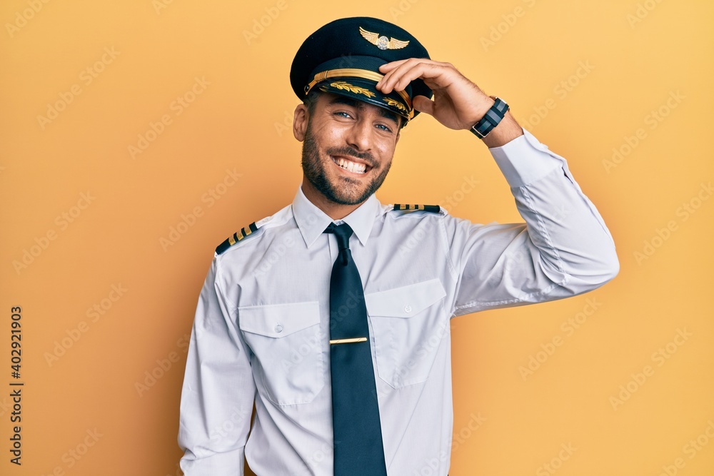 Handsome hispanic man wearing airplane pilot uniform smiling confident touching hair with hand up gesture, posing attractive and fashionable - obrazy, fototapety, plakaty 