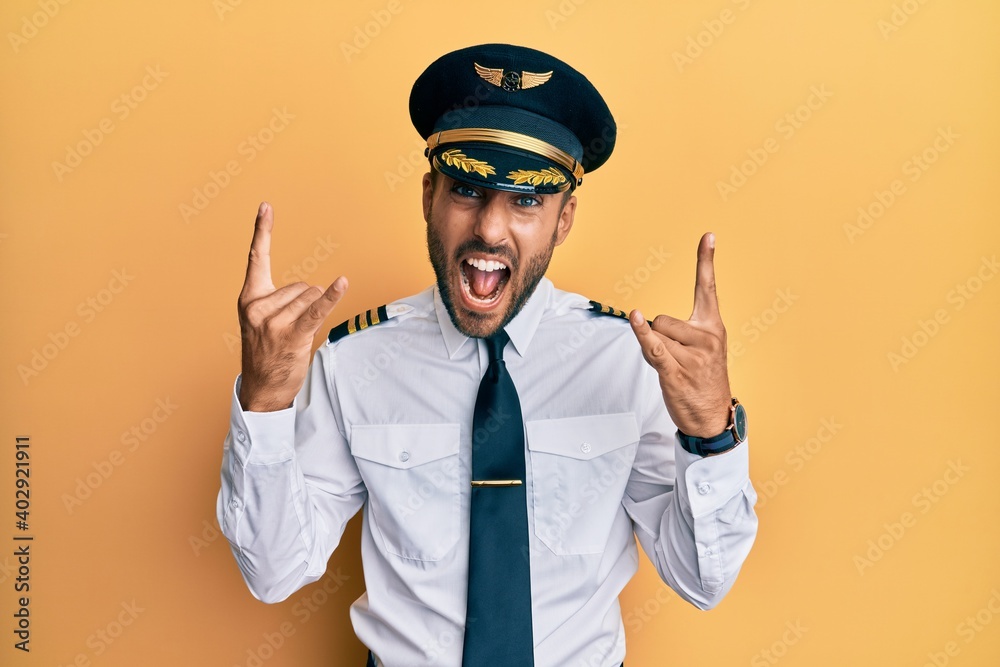 Handsome hispanic man wearing airplane pilot uniform shouting with crazy expression doing rock symbol with hands up. music star. heavy music concept. Handsome hispanic man wearing airplane pilot uniform shouting with crazy expression doing rock symbol with hands up. music star. heavy music concept.