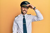 Handsome hispanic man wearing airplane pilot uniform smiling confident touching hair with hand up gesture, posing attractive and fashionable