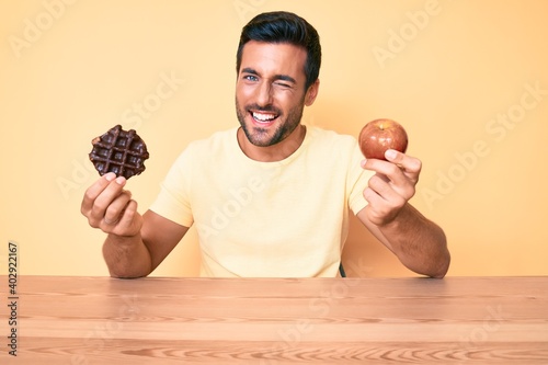 Young hispanic man eating breakfast holding chocolate waflle and apple winking looking at the camera with sexy expression, cheerful and happy face. photo