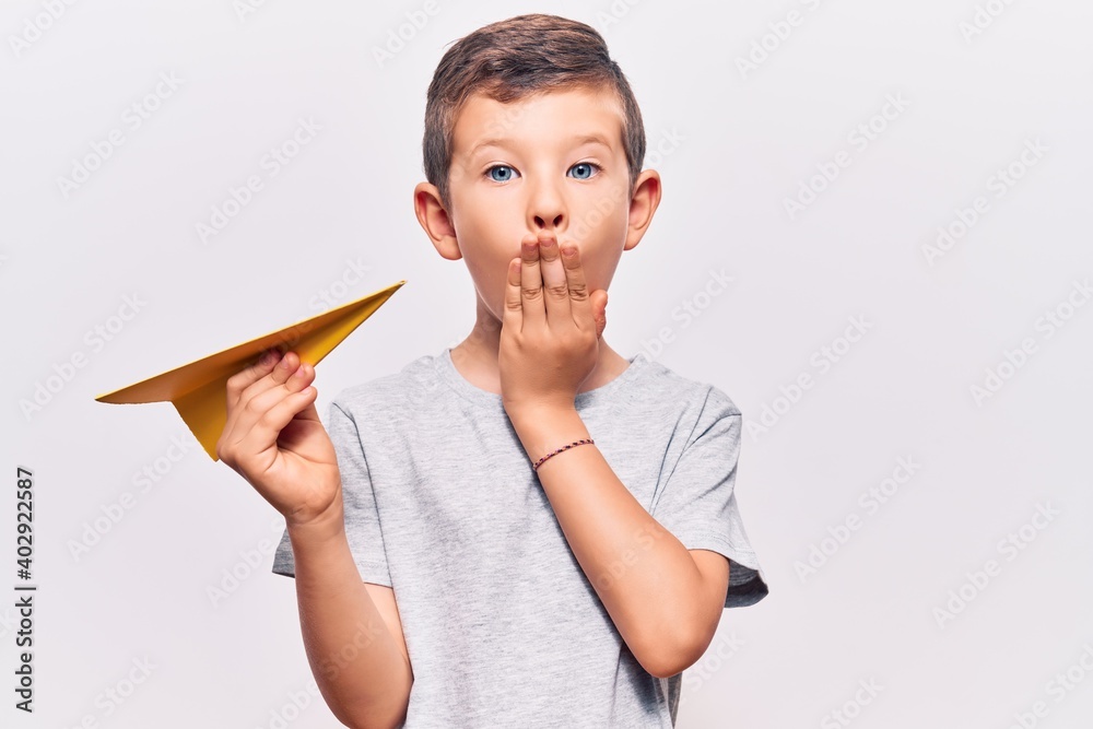 Cute blond kid holding paper airplane covering mouth with hand, shocked and afraid for mistake. surprised expression Cute blond kid holding paper airplane covering mouth with hand, shocked and afraid for mistake. surprised expression