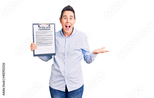 Young woman with short hair holding clipboard with terms and conditions document celebrating victory with happy smile and winner expression with raised hands