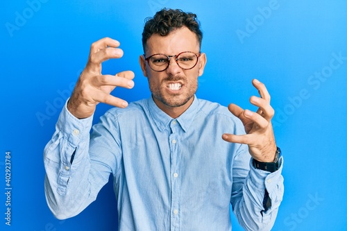 Young hispanic man wearing casual clothes and glasses shouting frustrated with rage, hands trying to strangle, yelling mad