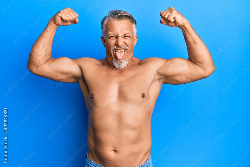Middle age grey-haired man standing shirtless doing fitness gesture sticking tongue out happy with funny expression.
