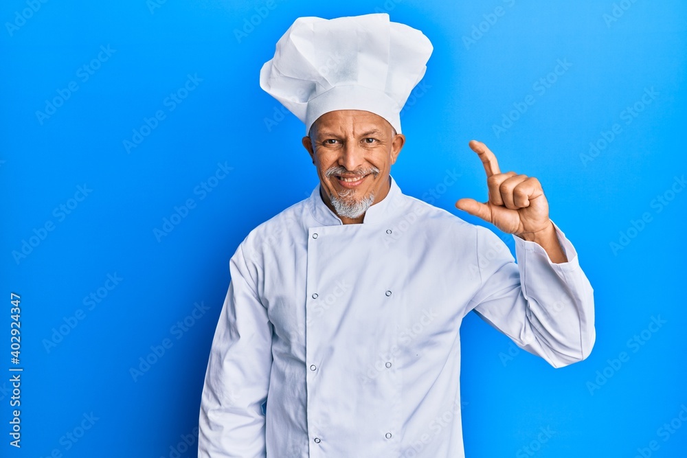 Middle age grey-haired man wearing professional cook uniform and hat smiling and confident gesturing with hand doing small size sign with fingers looking and the camera. measure concept. Middle age grey-haired man wearing professional cook uniform and hat smiling and confident gesturing with hand doing small size sign with fingers looking and the camera. measure concept.