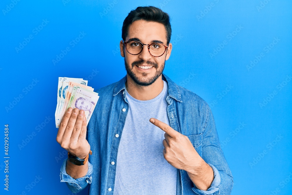 Young hispanic man holding singapore dollars banknotes smiling happy pointing with hand and finger Young hispanic man holding singapore dollars banknotes smiling happy pointing with hand and finger