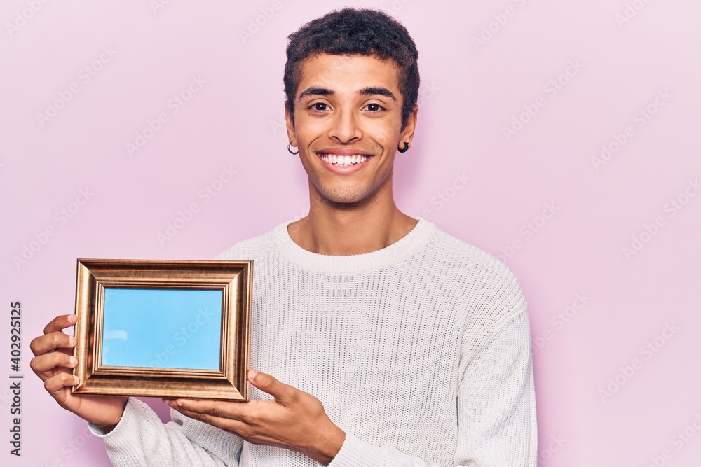 Young african amercian man holding empty frame looking positive and happy standing and smiling with a confident smile showing teeth