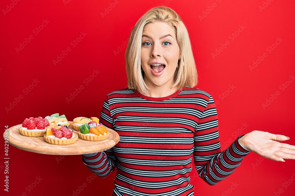 Young blonde woman holding sweet pastries celebrating achievement with happy smile and winner expression with raised hand