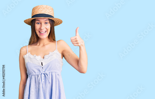 Beautiful brunette young woman wearing summer hat smiling happy and positive, thumb up doing excellent and approval sign