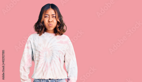 Young beautiful mixed race woman wearing casual tie dye sweatshirt puffing cheeks with funny face. mouth inflated with air, crazy expression.