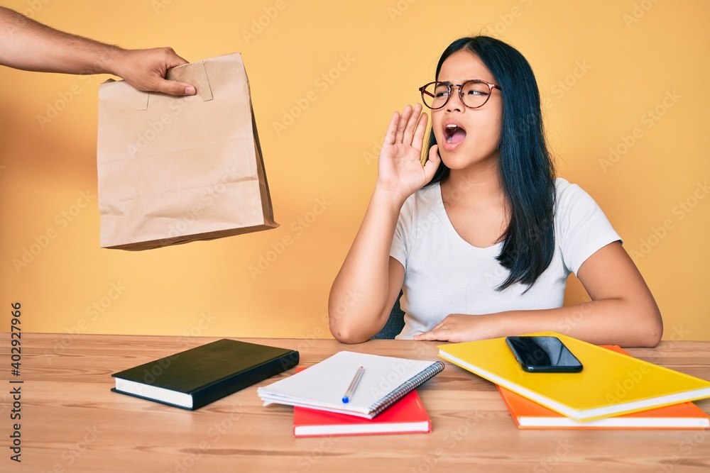 Young beautiful asian girl sitting on the table stuying getting take away food shouting and screaming loud to side with hand on mouth. communication concept. Young beautiful asian girl sitting on the table stuying getting take away food shouting and screaming loud to side with hand on mouth. communication concept.
