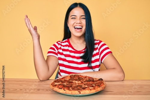 Young beautiful asian girl eating tasty pepperoni pizza celebrating victory with happy smile and winner expression with raised hands