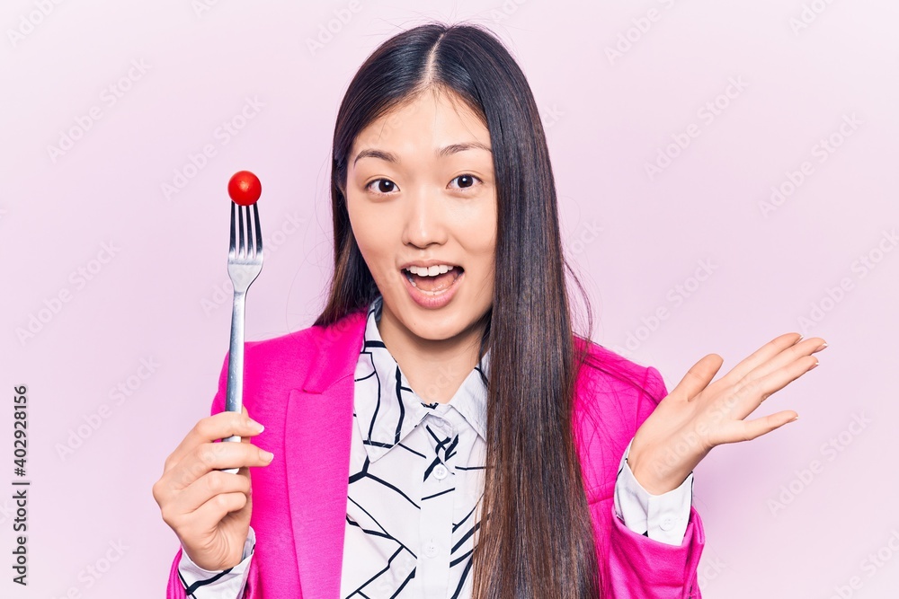 Young beautiful chinese woman holding fork with tomato celebrating achievement with happy smile and winner expression with raised hand Young beautiful chinese woman holding fork with tomato celebrating achievement with happy smile and winner expression with raised hand