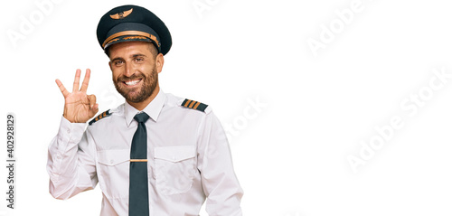 Handsome man with beard wearing airplane pilot uniform smiling positive doing ok sign with hand and fingers. successful expression.