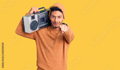 Handsome latin american young man holding boombox, listening to music pointing to you and the camera with fingers, smiling positive and cheerful