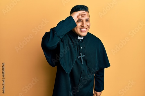 Young latin priest man standing over yellow background very happy and smiling looking far away with hand over head. searching concept.