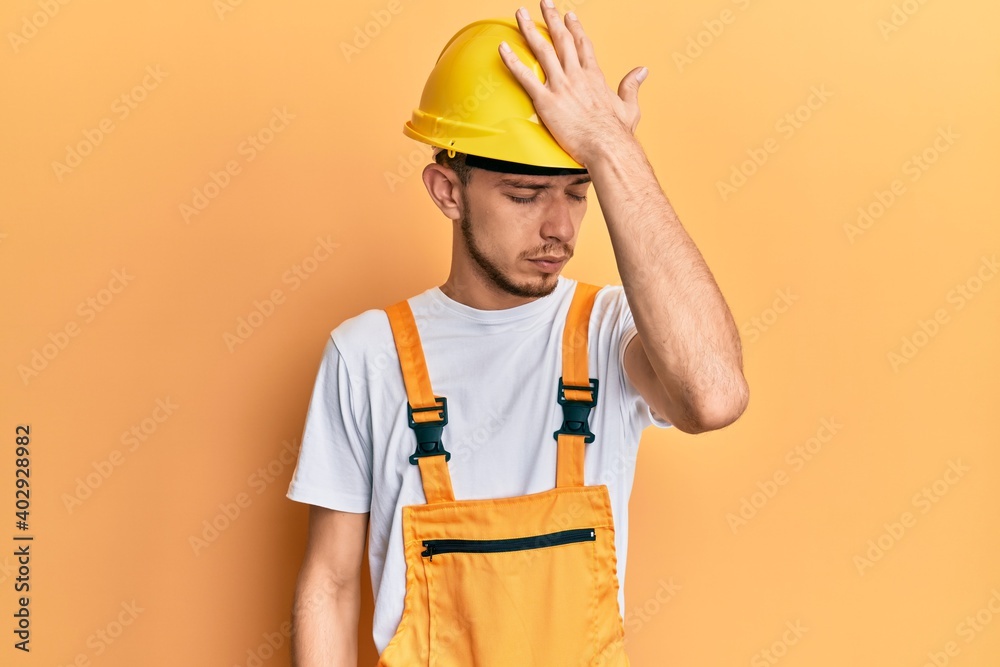 Hispanic young man wearing builder uniform and safety hardhat surprised with hand on head for mistake, remember error. forgot, bad memory concept. Hispanic young man wearing builder uniform and safety hardhat surprised with hand on head for mistake, remember error. forgot, bad memory concept.