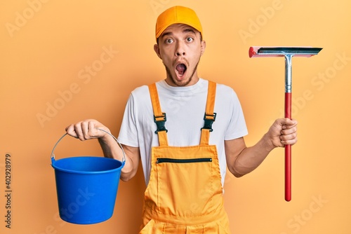Photography Hispanic young man wearing glass cleaner uniform and squeegee afraid and shocked with surprise and amazed expression, fear and excited face