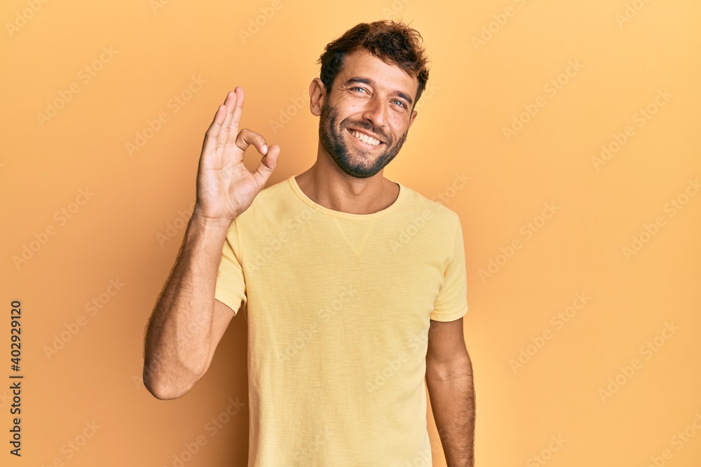 Handsome man with beard wearing casual yellow tshirt over yellow background smiling positive doing ok sign with hand and fingers. successful expression. Handsome man with beard wearing casual yellow tshirt over yellow background smiling positive doing ok sign with hand and fingers. successful expression.
